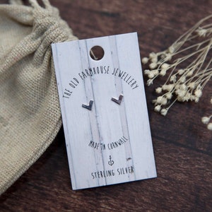 May include: A pair of sterling silver earrings with a small, angled heart shape. The earrings are displayed on a white card with the text "The Old Farmhouse Jewellery" and "Made in Cornwall" printed on it.