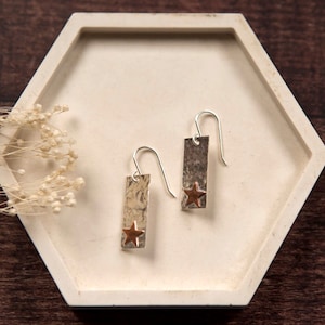 May include: Silver dangle earrings with a hammered rectangular shape and a copper star charm.