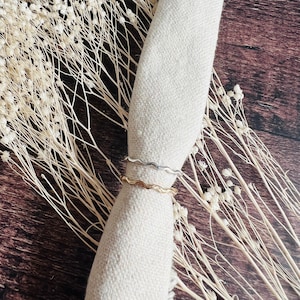 May include: Two wavy rings, one gold and one silver, are stacked on top of each other on a white fabric background.