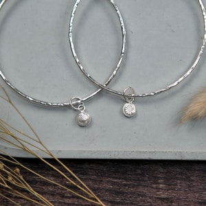 May include: Two silver bangles with hammered textures and small silver charms. The charms are round and have a floral design.