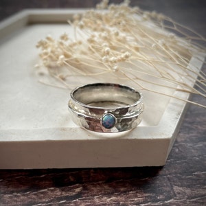 May include: A silver ring with a hammered texture and a small, round, blue opal gemstone. The ring is displayed on a white tray. The ring's design features a central stone and a textured band. The background includes dried flowers.