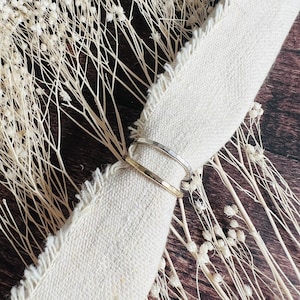 May include: Two hammered metal rings, one silver and one gold, are stacked on a piece of white fabric. The fabric is on a wooden surface with dried white flowers.