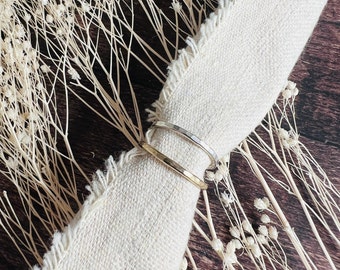 May include: Two hammered metal rings, one silver and one gold, are stacked on a piece of white fabric. The fabric is on a wooden surface with dried white flowers.