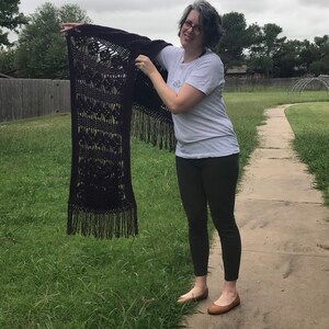May include: A woman is holding a dark purple crocheted shawl with fringe. The shawl has a geometric pattern and is made with a fine yarn.