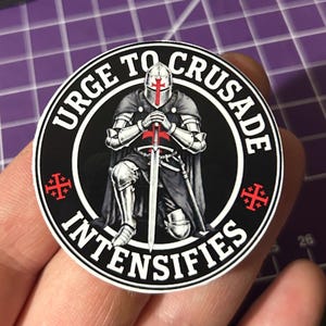 Puede incluir: Una pegatina circular que muestra a un caballero con armadura plateada arrodillado con una espada, usando un casco con una cruz roja. El borde negro contiene el texto blanco "URGE TO CRUSADE INTENSIFIES" y dos pequeñas cruces rojas laterales.