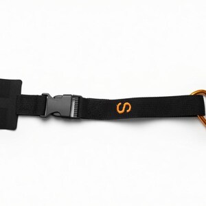 Peut inclure: Une sangle en nylon noir dotée d'une extrémité en tissu rectangulaire, d'une boucle en plastique à ouverture latérale et d'une lettre S brodée en orange. La sangle comprend un mousqueton orange métallique fixé à une extrémité pour la fixation.