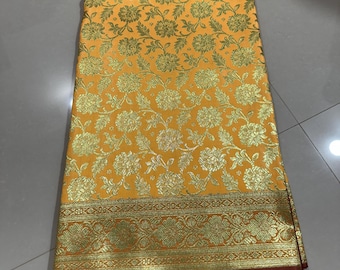 May include: A folded fabric textile featuring a vibrant yellow base with an intricate gold floral pattern throughout. The bottom edge displays a wide, ornate gold border with detailed botanical motifs, finished with a deep maroon hemline.
