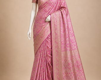 May include: A pink sari featuring intricate gold metallic floral and paisley patterns throughout the fabric. The garment includes a wide, ornate gold border with detailed motifs and a matching short-sleeved blouse. The text VATANI SARI is visible.