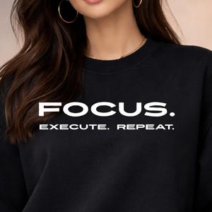 Könnte beinhalten: Schwarzes Sweatshirt mit Rundhalsausschnitt und dem weißen Text „FOCUS.“ und „EXECUTE. REPEAT.“ Das Sweatshirt ist einfarbig schwarz und hat einen klassischen, lässigen Stil. Der Text ist fett und ohne Serifen.