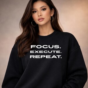 May include: Black crew neck sweatshirt with the words "FOCUS. EXECUTE. REPEAT." in white block letters. The sweatshirt is a solid black colour and has a classic, casual style.