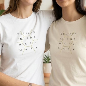 May include: Two white t-shirts with the text "BELIEVE IN THE HAIL MARY" in black and gold. The text is arranged in a hexagonal design. The shirts are plain and simple.