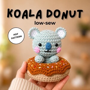 May include: A crochet koala sitting inside a brown donut with colorful sprinkles. The text KOALA DONUT low-sew and PDF PATTERN are visible. The koala has light blue yarn, a dark blue nose, black eyes, and pink cheeks on a round head.