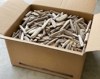May include: A large cardboard box filled with a collection of light gray and tan weathered driftwood pieces. The wooden fragments vary in length and thickness, featuring smooth, rounded edges and natural textures from exposure to water and sand.