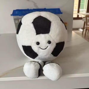 May include: A plush soccer ball with a smiling face and two small feet. The ball is primarily white with black pentagon-shaped patches, resembling a classic soccer ball design. The toy is soft and cuddly, perfect for children.