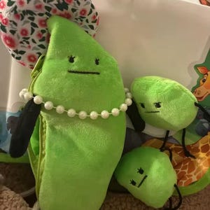 May include: A plush green pea pod with a pearl necklace and a floral hat, along with three smaller pea plushies. The pea pod has a zipper and black arms. The plushies have embroidered faces and black string legs.