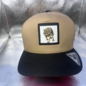 May include: A tan baseball cap with a black brim and a square patch on the front featuring a detailed illustration of a wolf. A black sticker on the brim displays the text 804 and Premium Quality Merchandise in white lettering.
