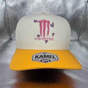 May include: A cream-colored baseball cap with a yellow brim. The front features an embroidered pink Monster logo surrounded by small purple hearts. A white sticker with black text reading KAMEL PREMIUM QUALITY HEADWEAR 804 is on the brim.