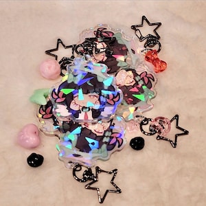Può includere: Una collezione di portachiavi di personaggi in stile anime con accenti olografici. I portachiavi presentano un personaggio in abito nero con cravatta rossa. Sono inclusi anche ciondoli a forma di stella, perline rosa a forma di cuore e perline nere.