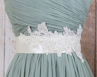 May include: A sage green dress with a pleated bodice and skirt. A white satin sash with delicate floral lace detailing accents the waist. The dress is likely for a special occasion, such as a wedding or formal event.