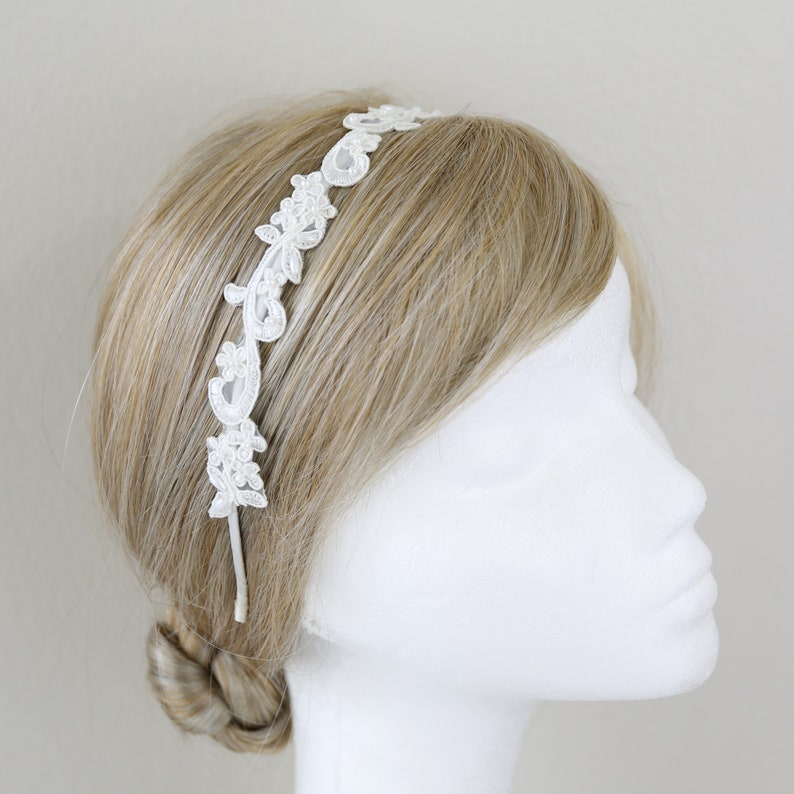 May include: White lace headband with floral and leaf designs. The headband is displayed on a mannequin head with blonde hair. The delicate lace detailing adds a touch of elegance, perfect for weddings or special occasions.