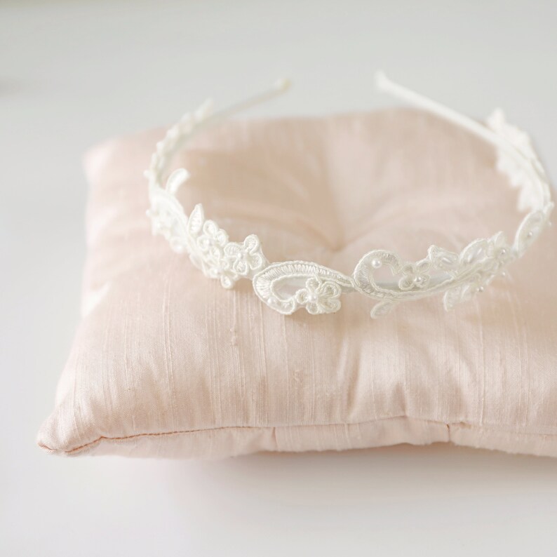 May include: A delicate white lace headband with floral and leaf designs, adorned with small pearls. The headband rests on a soft, light pink satin pillow, creating a romantic and elegant aesthetic. Perfect for weddings or special occasions.