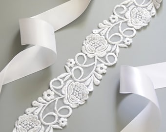 May include: White floral lace trim with a delicate design. The lace is on a white satin ribbon.