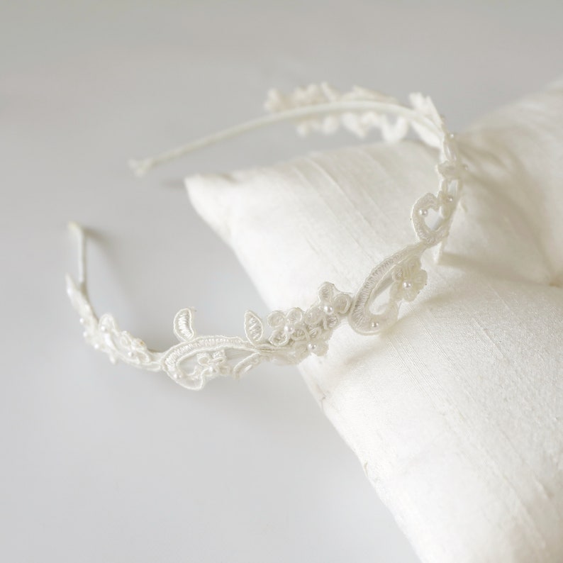 May include: A delicate, off-white bridal headband with floral and pearl accents. The headband is made of lace and sits atop a white satin pillow. The design features intricate floral patterns and small, round pearls.