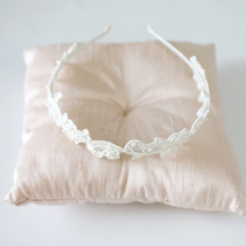 May include: A delicate white lace headband with floral and leaf details. The headband is set on a light pink satin pillow, creating a soft and elegant presentation. Perfect for weddings or special occasions.