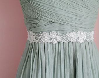 May include: A sage green dress featuring a bodice with horizontal pleats and a waistline accented by a white floral lace applique. The skirt consists of vertical pleats that fall from the waist, creating a textured and layered appearance.