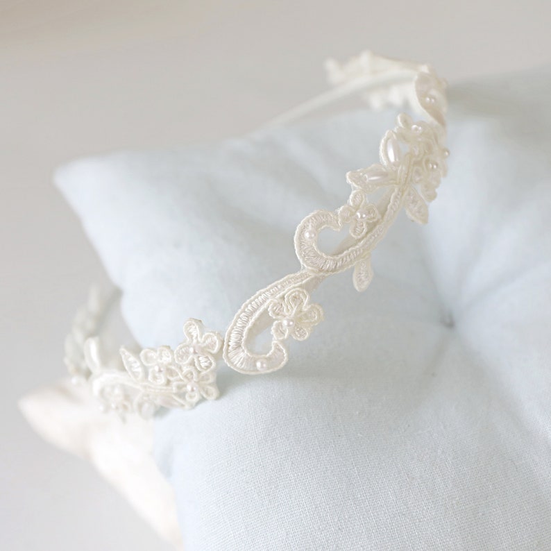 May include: Ivory-colored lace headband with floral and leaf designs. The delicate headpiece is adorned with small, pearl-like beads. The headband is resting on a light blue pillow, showcasing its intricate details.