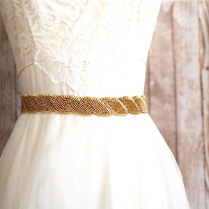 May include: A gold beaded belt featuring a repeating diagonal chevron pattern, worn around the waist of a white lace dress. The belt is constructed from small, metallic gold seed beads arranged in a dense, textured, and continuous braided design.