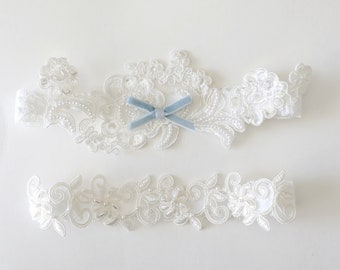 May include: Two-piece white lace wedding garter set. The top garter features floral embroidery, pearl accents, and a light blue velvet bow. The bottom garter has floral and leaf embroidery with pearl accents. Both are set against a white background.