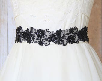 May include: A white dress with a black floral lace sash. The sash is beaded and has a delicate design.