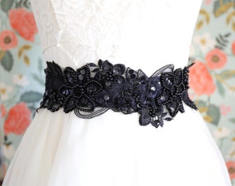 May include: A black lace belt featuring intricate floral embroidery, embellished with small black beads and sequins. The belt is worn around the waist of a white dress, showcasing a detailed pattern of petals and leaves across the dark fabric.