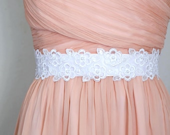 May include: A white floral lace sash with pearl accents is wrapped around a peach-colored dress. The sash is the focal point, with delicate flower patterns and a satin-like texture. The dress has a draped bodice and a flowing skirt.