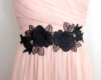 May include: A pink dress with a black lace floral sash. The sash is made of delicate black lace and features a floral design with leaves and flowers.
