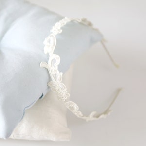 May include: Ivory lace headband with floral and leaf details. The delicate design features small, pearl-like embellishments. The headband is set against a light blue and white background, suggesting a bridal or special occasion theme.