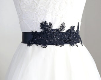 May include: A black satin sash belt featuring intricate black lace floral embroidery, delicate beadwork, and sequins. The belt is worn around the waist of a white lace dress, showcasing a detailed botanical pattern with dark embellishments.