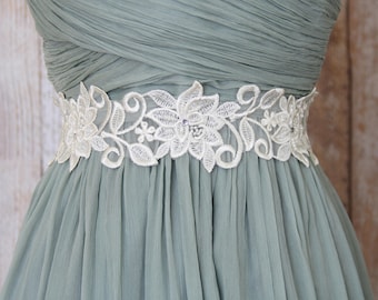 May include: A sage green dress with a white floral lace belt. The belt features intricate floral and leaf designs with small, sparkling embellishments. The dress has a pleated bodice and a flowing skirt, creating an elegant silhouette.
