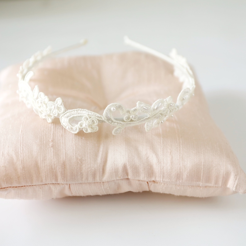 May include: A delicate, white lace headband with floral and leaf designs. The headband is adorned with small, subtle embellishments and rests on a light pink satin pillow. Perfect for weddings or special occasions.