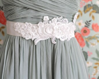 May include: A sage green dress with a white floral lace belt. The dress has a pleated bodice and skirt. The belt features intricate white lace and pearl accents, adding a touch of elegance to the dress.
