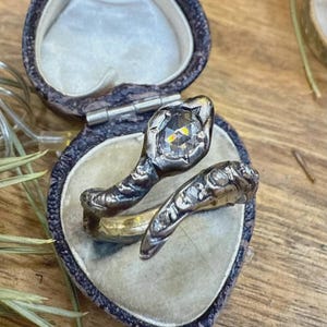 Op de afbeelding: Een vintage slangenring met een grote, facetgeslepen edelsteen op de kop, gezet in donker metaal. De ring wordt getoond in een hartvormige sieradendoos met een fluwelen binnenkant, die het ingewikkelde ontwerp en de antieke esthetiek benadrukt.