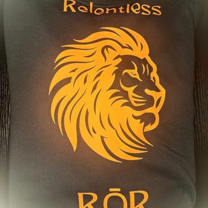 May include: Dark grey t-shirt featuring an orange lion's head design. The word "Relentless" is printed above the lion, and the word "Rōr" is printed below. The t-shirt is folded.