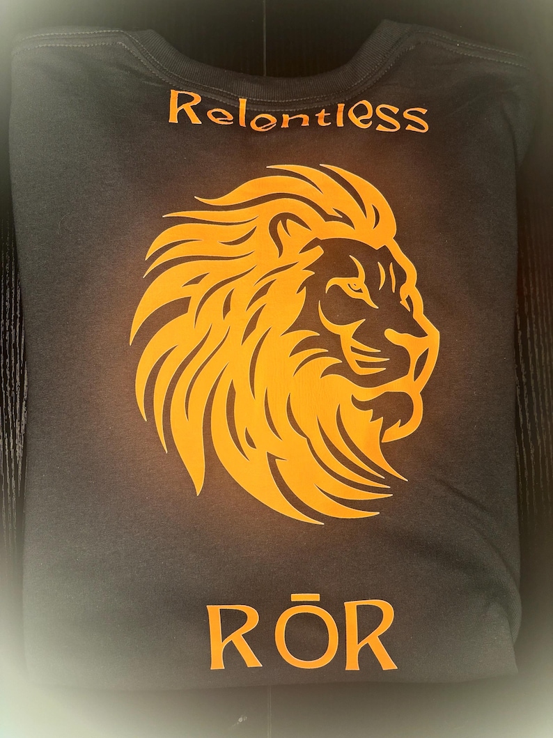 May include: Dark grey t-shirt featuring an orange lion's head design. The word "Relentless" is printed above the lion, and the word "Rōr" is printed below. The t-shirt is folded.