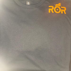 May include: Dark grey crew neck t-shirt featuring the orange text "ROR" and a lion graphic. The shirt is made of a soft material. The brand tag "DAN" is visible on the collar.