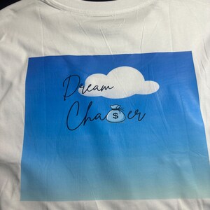 May include: White t-shirt with a blue gradient graphic. The graphic features a cloud, the words "Dream Chaser" in cursive, and a money bag icon.
