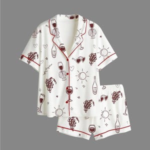Può includere: Pigiama bianco con stampa a tema vino. La maglia a maniche corte e i pantaloncini presentano un motivo di calici, bottiglie, uva e cuori, delineati in rosso scuro. Il set ha finiture e bottoni rosso scuro.
