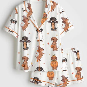 May include: A white short-sleeved button-down pajama shirt and matching shorts set featuring an all-over print of dachshunds dressed in Halloween costumes, including pumpkins, witch hats, and bat wings, accented with orange piping trim.