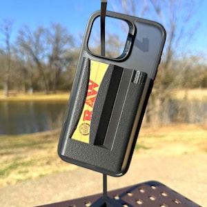 Puede incluir: Funda de teléfono negra con un compartimento incorporado que contiene un paquete de papel RAW amarillo y rojo. La funda está montada sobre un soporte metálico sobre un fondo exterior borroso. La funda está diseñada para guardar un teléfono y accesorios.