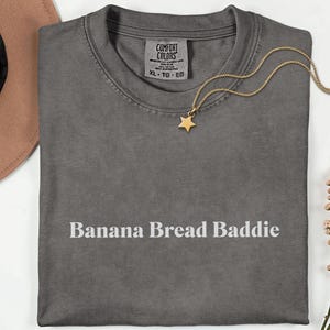 May include: A grey Comfort Colors t-shirt with the text "Banana Bread Baddie" in white. A gold star pendant necklace and a brown hat are also visible. The shirt is folded neatly.